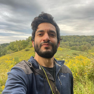 person with short hair and a beard wearing a jacket on a forested hill