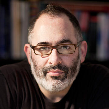 person with beard and glasses and a black t-shirt in front of a bookcase