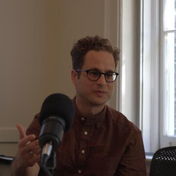 person with glasses wearing a brown button down shirt speaking into a microphone